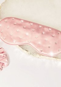 Różowa satynowa maska do spania ozdobiona małymi, przezroczystymi kryształkami, o gładkiej fakturze i wyprofilowanym kształcie, blokującym światło. Obok znajduje się pasująca gumka do włosów.