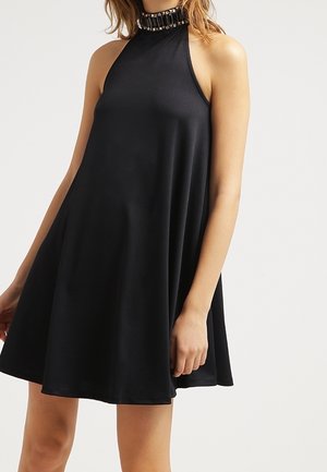 Woman wearing sleeveless black A-line dress with high neckline and decorative beaded collar against plain light background.