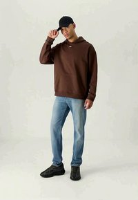 Sweatshirt castanho com um bolso à frente, combinado com jeans azul claros e ténis pretos. O modelo usa um boné preto e está de pé contra um fundo claro.