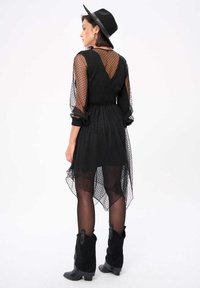 Zwart stippen netwerkjurk met lange doorschijnende mouwen, gerimpelde taille en een asymmetrische zoom, gecombineerd met zwarte enkellaarsjes met cowboy-inspiratie en een zwarte hoed.