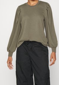 Jersey verde oliva con mangas largas y abullonadas, y un escote redondo, combinado con pantalones cargo negros. Textura suave, corte relajado.