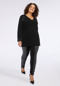 Bluza neagră cu mânecă lungă, cu accente cu mărgele și decolteu în V, asortată cu colanți negri lucioși și pantofi cu toc înalt negri.