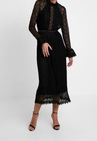 Robe en dentelle noire avec col montant, texture plissée et ourlet festonné. Accentuée par de longues manches avec poignets et une ceinture en tissu.