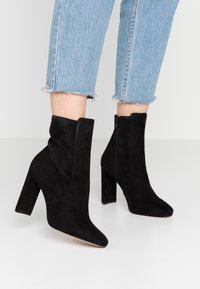 Bottes chevilles en daim noir avec bout pointu et talon bloc haut. Dotées d'une fermeture éclair latérale et d'une texture lisse, portées avec un jean bleu clair.