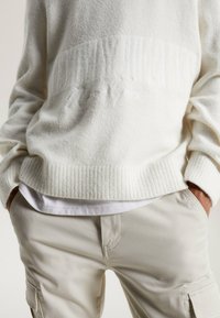 Krämfärgad tröja i mjuk stickad tyg med ribbade ärmslut och nederkant, med diskret broderi som säger "New York" framtill, i kombination med beige byxor.