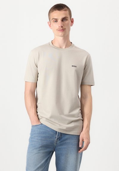 T-shirt en coton beige clair à manches courtes et col rond. Présente un petit logo noir sur la poitrine gauche. Porté avec un jean bleu.