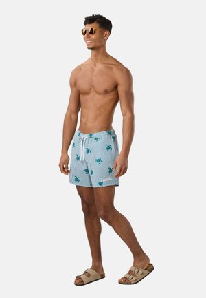 Muž bez košele, ktorý má na sebe vzorované plavkové šortky, béžové sandále a slnečné okuliare, stojí v uvoľnenej, usmiatej póze na bielom pozadí.