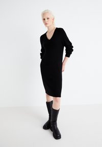 Black knit dress with a V-neck and long, cuffed sleeves, paired with knee-high black boots. Smooth texture and form-fitting design.