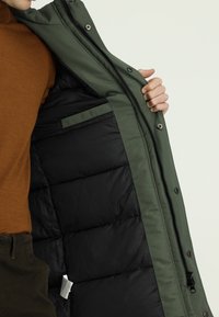 Osoba w brązowej koszuli trzyma otwartą zieloną kurtkę, pokazując jej pikowaną czarną podszewkę i wewnętrzną kieszeń.