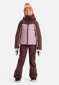Paarse en kastanjebruine winterjas met een rits, gecombineerd met bijpassende broek. Oranje muts met zwarte letterdruk en gemêleerde handschoenen. Zwarte laarzen.