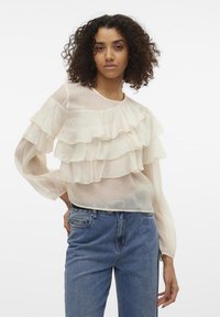 Blouse beige clair en tissu transparent, avec trois couches froncées à l'avant et des manches longues légèrement bouffantes.