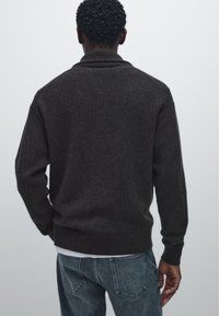 Donkerbruin gebreid pullover met een opstaande kraag, geribbelde manchetten en zoom. Draag het over een licht shirt, gecombineerd met grijze jeans, gezien vanuit de achterkant.
