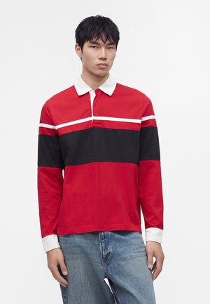 Jeune homme portant un polo à manches longues rouge et noir avec col et poignets blancs, associé à un jean bleu, sur fond uni.