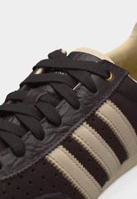 Σκούρο καφέ και μπεζ αθλητικό παπούτσι με μαύρα κορδόνια, διάτρητα πλευρικά πάνελ και τρεις μπεζ ρίγες στο σκούρο καφέ δέρμα της επάνω επιφάνειας.