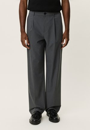 Pantalon gris sur mesure avec une coupe ample, doubles plis et fermeture par bouton. Confectionné dans un tissu texturé. Porté avec des chaussures noires.
