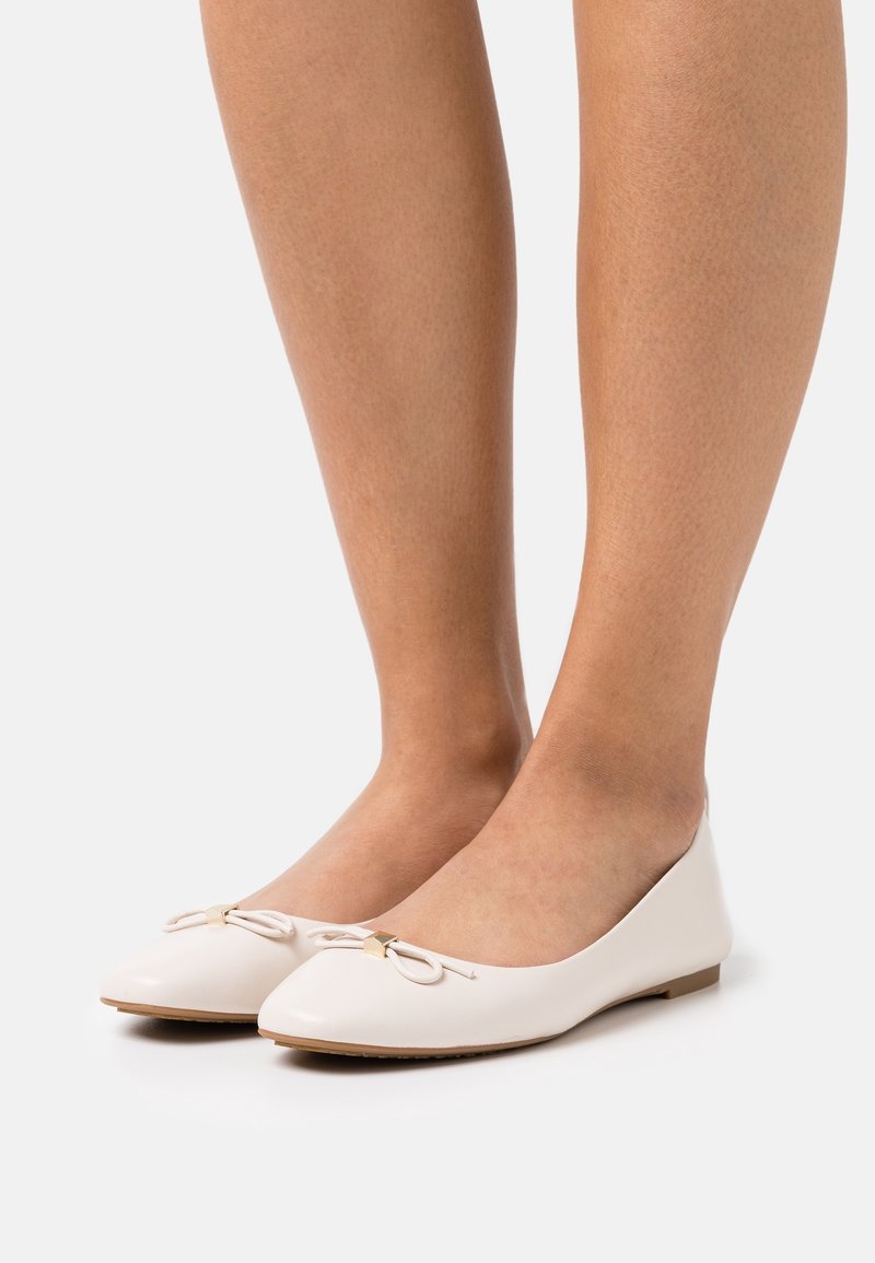 Jambes portant des ballerines blanches avec petits nœuds et touches dorées, debout sur un fond clair uni.