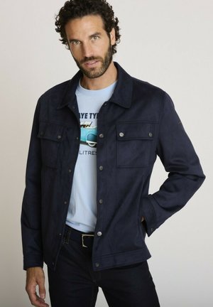 Homme aux cheveux bruns bouclés portant une veste bleu marine boutonnée par-dessus un t-shirt graphique bleu clair, debout avec une main dans la poche sur un fond uni.