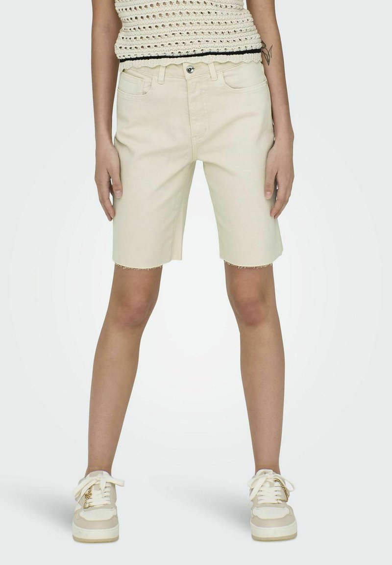 Shorts de denim beige claro con un dobladillo deshilachado, cinco bolsillos y un cierre delantero con botón, combinados con zapatillas blancas y una blusa de punto.