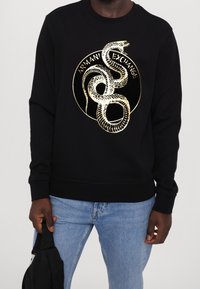 Svart sweatshirt med guldormgrafik och "Armani Exchange"-text, bärs med ljusblå jeans, person som håller en svart väska.
