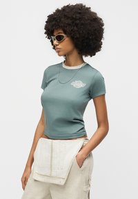 Γυναίκα με μεγάλα σγουρά μαλλιά, φοράει κορυφή με κοντά μανίκια σε τιρκουάζ χρώμα, μπεζ παντελόνι, γυαλιά ηλίου και αλυσίδα στο λαιμό, ποζάροντας με το ένα χέρι στην τσέπη.