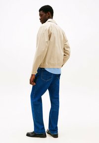 Beige lichte jas met een kraag, gecombineerd met een blauw gestreept shirt en blauwe denim jeans, gedragen met zwarte schoenen.