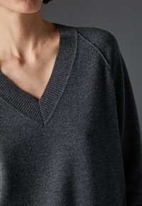 Siva v-neck pulover iz mehkega materiala z rebrastim ovratnikom in sproščenim krojem. Ima teksturirano površino brez vzorcev ali okraskov.