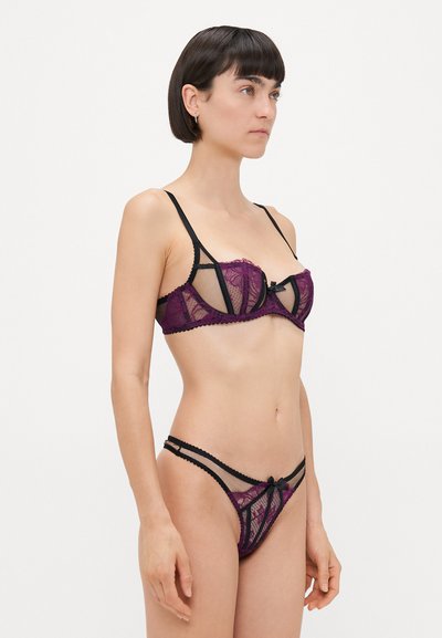 Set di lingerie in pizzo viola con cinturini neri. Caratterizzato da un design trasparente, motivi floreali e un accento a fiocco sia sul reggiseno che sul perizoma.