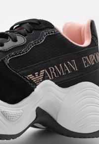 Svart atletisk sko med mocka- och mesh-material, med en rosa accent vid hälen, vit texturerad sula och en logotyp på sidan.