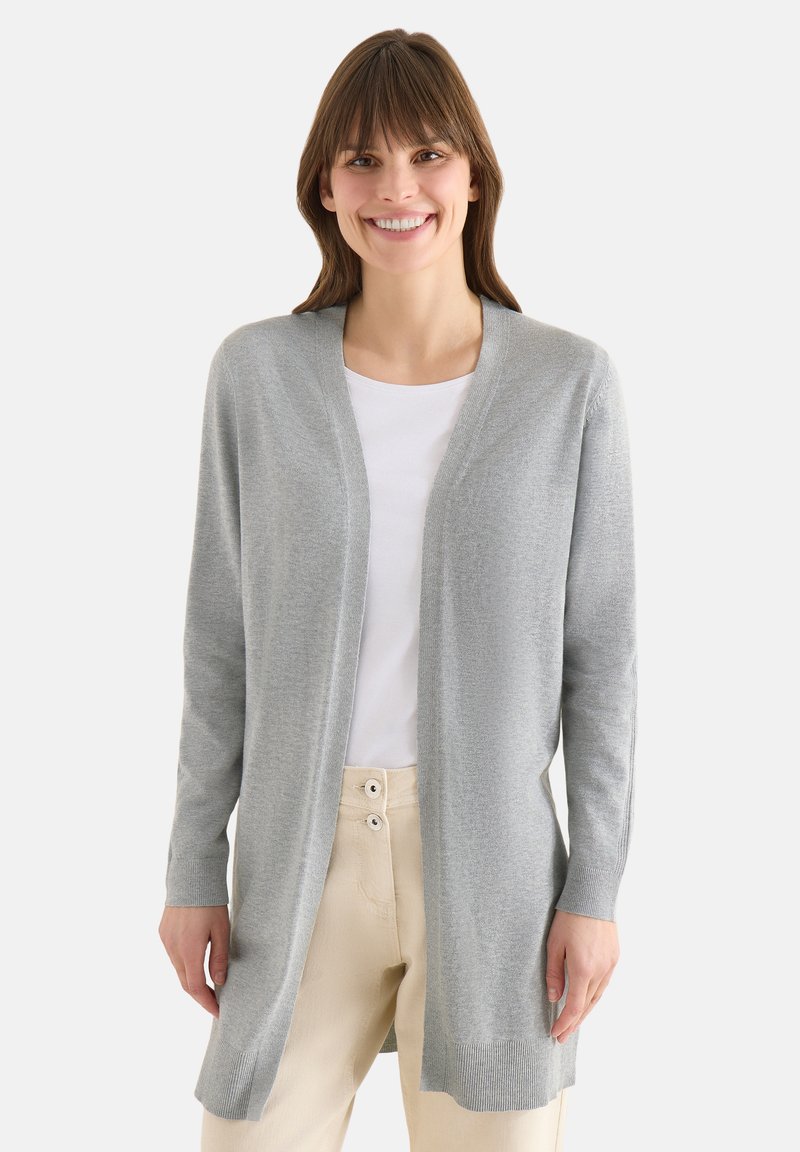 Femme souriante portant un cardigan gris clair ouvert sur un chemisier blanc et un pantalon beige, sur un fond clair uni.