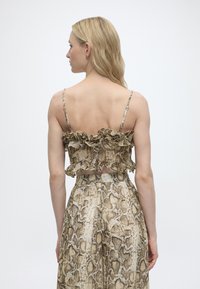 Ξανθιά γυναίκα με θέα από πίσω, φορώντας τοπ cropped με βολάν και σχέδιο φίδι, λεπτές τιράντες και ταιριαστό ψηλόμεσο παντελόνι, μπροστά σε απλό φόντο.