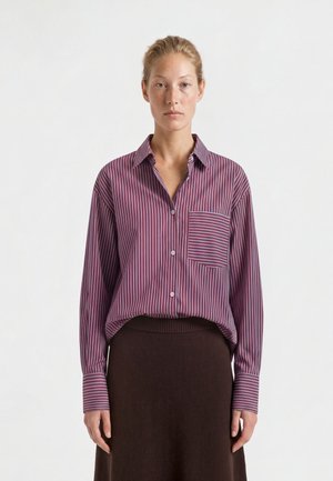 Femme aux cheveux blonds portant une chemise ample rayée violette et blanche, rentrée dans une jupe marron foncé, debout devant un fond uni.