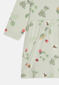Lys grøn kjole i stoffet med et blomsterdesign som har bier, marihøner og blader. Lange ermer med en samlet midje detalj.