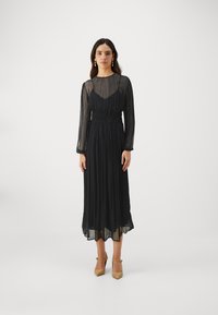 Sort plisseret kjole med lange gennemsigtige ærmer og en tætsiddende talje. Inkluderer en mellemlang nederdel og en rundet udskæring. Bæres med beige hæle.