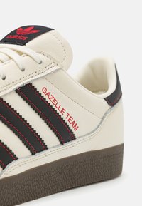 Sneaker Adidas Gazelle Team charakteryzuje się kremową skórą, trzema czarnymi paskami, czerwonymi akcentami oraz teksturowaną brązową gumową podeszwą.