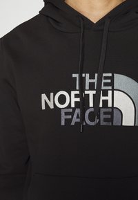 Gros plan sur un sweat à capuche noir avec le logo "THE NORTH FACE" et l'emblème demi-dôme en blanc et gris sur la poitrine.