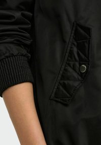 Veste bomber noire avec des détails de poche matelassée, des poignets côtelés et un tissu en nylon lisse. La vue en gros plan met en évidence la texture et le design.