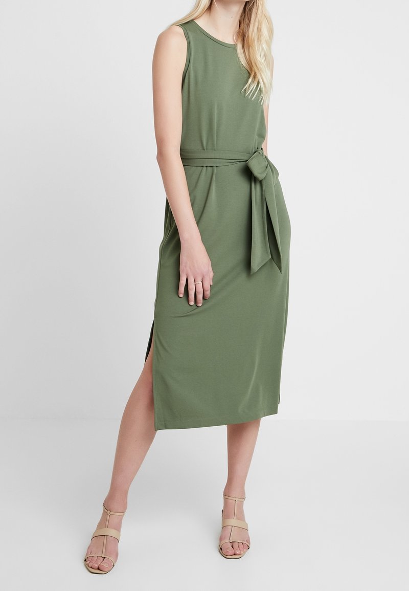 Abito senza maniche di colore verde oliva con una cintura in vita e uno spacco laterale, realizzato in un tessuto morbido. Abbinato a sandali strappy color beige chiaro.