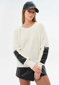 White knit sweater with black faux leather accents on sleeves and hem. Long sleeves and ribbed texture. Black cap. Model poses with hands crossed.