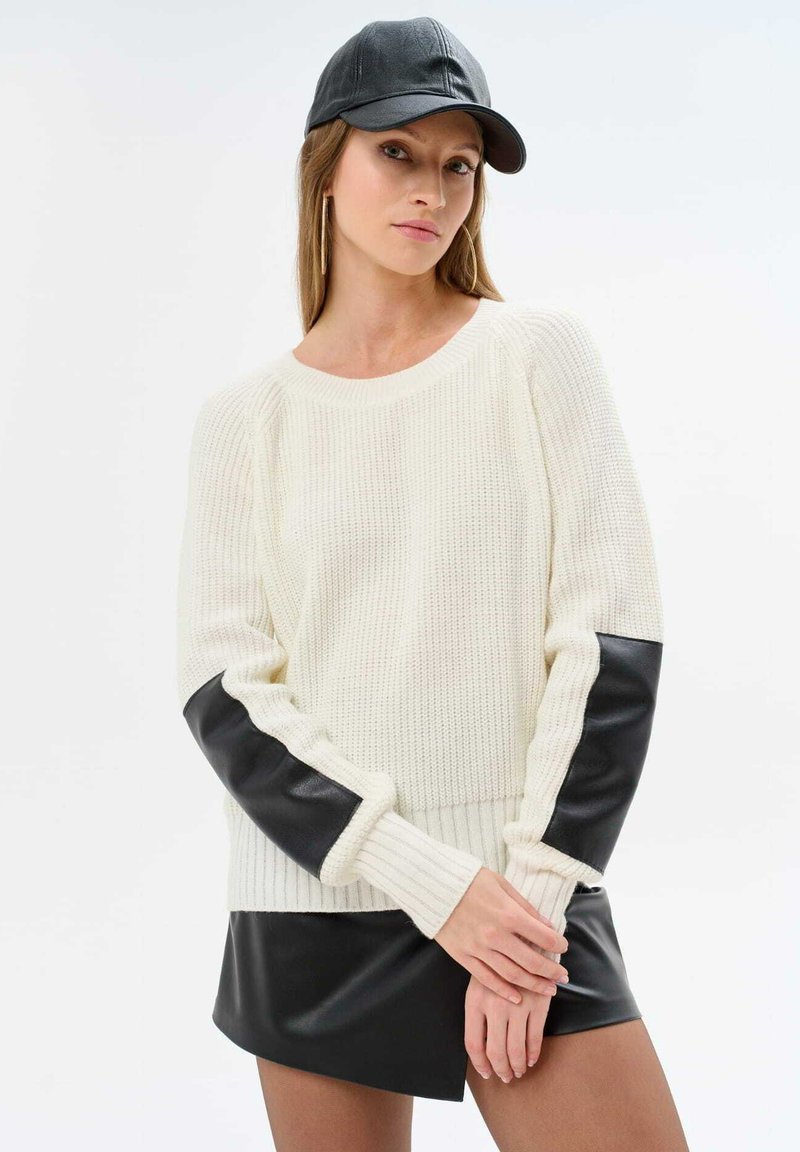 White knit sweater with black faux leather accents on sleeves and hem. Long sleeves and ribbed texture. Black cap. Model poses with hands crossed.