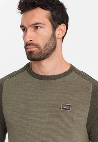 Maglione a coste verde oliva con pannelli spalla testurizzati a contrasto. Presenta una piccola etichetta con il logo sul petto. Collo rotondo.