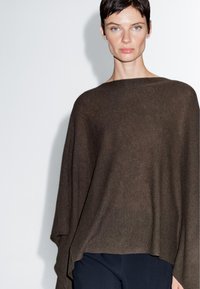 Brauner gestrickter Poncho mit weitem Ausschnitt, fließender Silhouette und strukturiertem Stoff, kombiniert mit dunklen Hosen vor einem weißen Hintergrund.