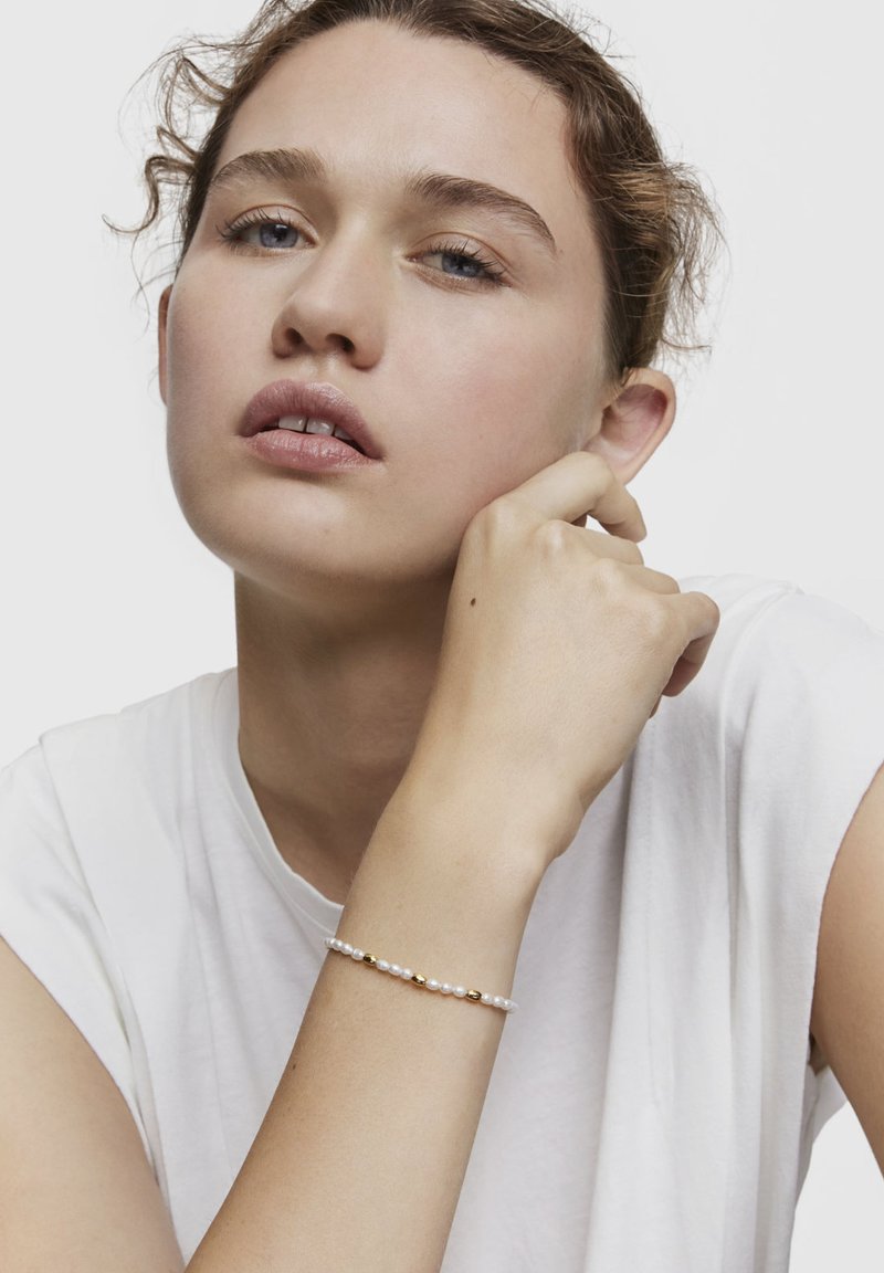 Giovane donna dai capelli ricci con indosso una maglietta bianca, poggiando il mento sulla mano, con un delicato braccialetto di perle e perline dorate su sfondo chiaro.