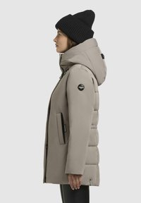 Polstret grå parka med høj krave, lynlåslommer og et sort logo-patch. Båret med en sort ribbet beanie. Sideprofil.