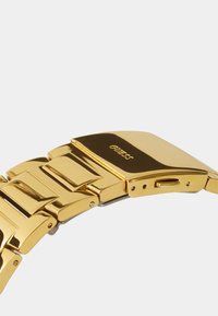 Západka a náramok hodiniek zo zlatého kovu s lešteným povrchom a vyrytým názvom značky „GUESS“ na západke.