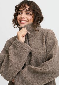 Brun fleece-pullover med høj krave, lynlås og oversized ærmer. Blød tekstur med et hyggeligt, varmt udseende. Detaljer omfatter en frontlomme.