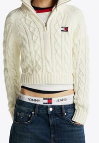 Femme portant un pull court blanc en maille torsadée, un jean bleu, avec la ceinture Tommy Jeans visible ainsi que le logo sur le pull et la poche du jean.
