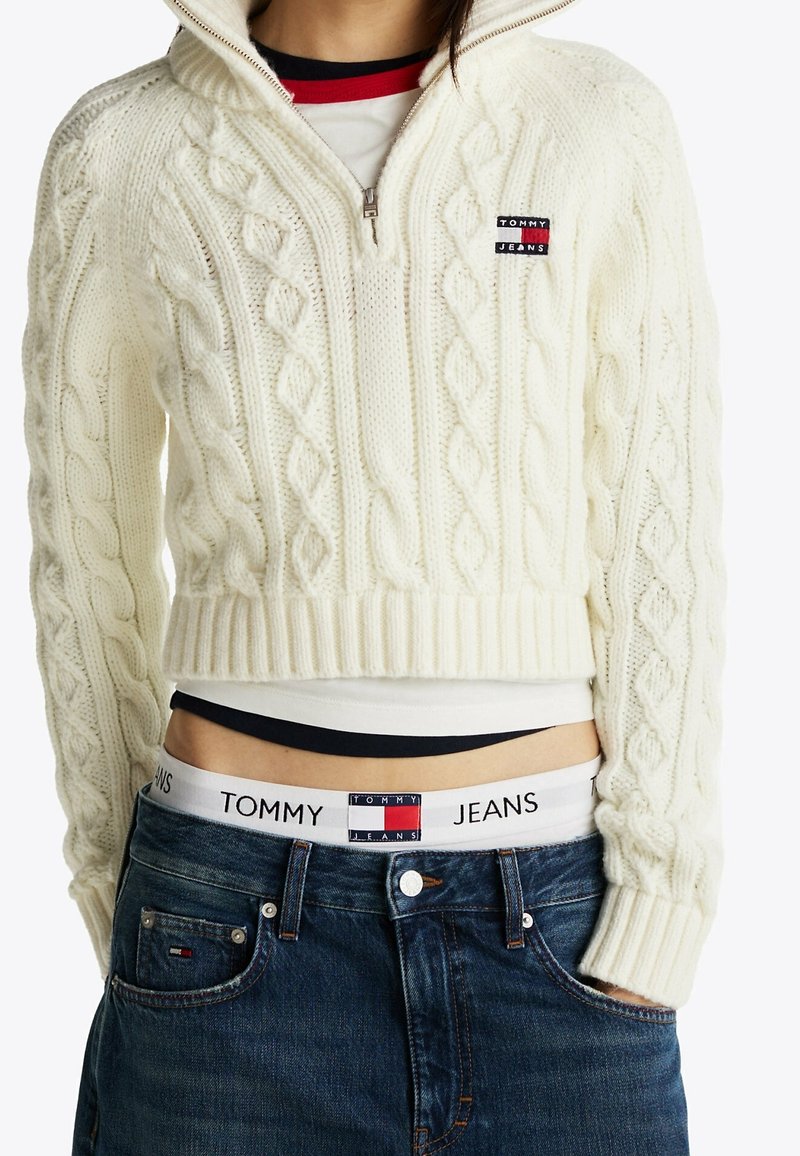 Femme portant un pull court blanc en maille torsadée, un jean bleu, avec la ceinture Tommy Jeans visible ainsi que le logo sur le pull et la poche du jean.