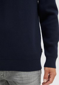 Marineblauwe gebreide trui met geribde manchetten en onderkant, gedragen over lichtgrijze jeans. De stof vertoont een gestructureerd patroon, zonder zichtbare logo's of prints.