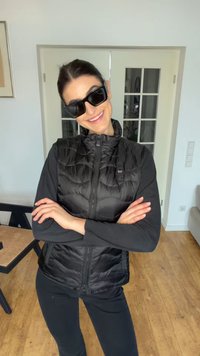 Donna al chiuso che sorride, indossa occhiali da sole neri, un gilet nero trapuntato sopra una camicia a maniche lunghe, con le braccia incrociate davanti a uno sfondo di pavimento in legno.