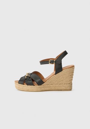 Sandale compensée en cuir noir avec semelle en jute tissée, bride de cheville avec boucle dorée et accent décoratif doré sur les brides avant.
